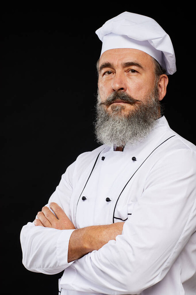 Retrato profesional de la cintura hacia arriba del chef sénior barbudo posando sobre fondo negro de pie con los brazos cruzados
 - Foto, imagen