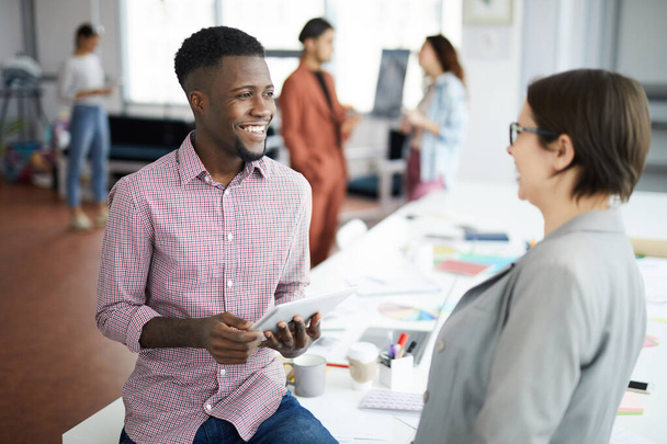 Portrait of smiling African-American man talking to female colleague while chatting casually in modern office, copy space - Foto, Imagem