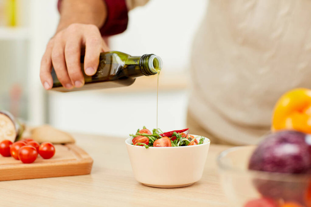 Close up of unrecognizable man pouring olive oil over healthy vegetable salad while cooking in kitchen, copy space - Foto, Bild