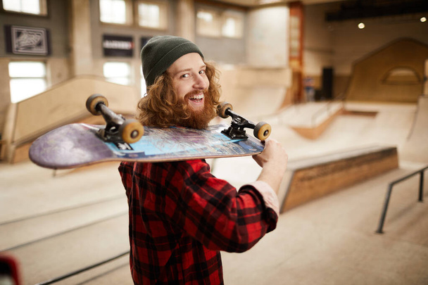 Cintura para cima retrato de hipster skater olhando para câmera sobre ombro enquanto posando no parque de patinação, espaço cópia
 - Foto, Imagem