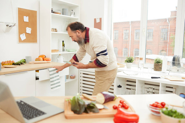 Retrato de vista lateral de hombre mayor barbudo abriendo cajones mientras cocina en la cocina, espacio para copiar
 - Foto, imagen