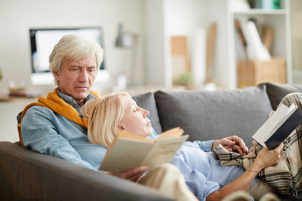 Retrato de pareja mayor moderna leyendo libros mientras está acostado en un cómodo sofá en casa, espacio para copiar
 - Foto, imagen