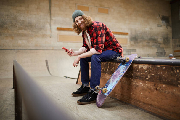 Full length portrait of contemporary bearded man looking at camera and smiling while using smartphone siting on ramp in skating park, copy space - Foto, immagini