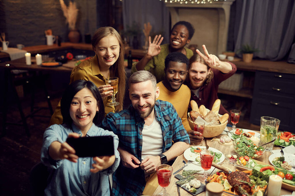 Multi-ethnic group of young friends taking selfie photo via smartphone and smiling happily while enjoying dinner party at home, copy space - 写真・画像