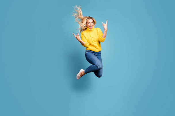 Full body photo of crazy lady jumping high enjoy metal rock concert showing hands horns excited wear knitted yellow pullover jeans isolated blue color background - Foto, afbeelding