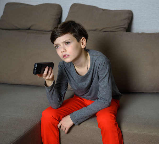 Concept boy teenager watching TV at home sitting on a sofa with different emotions. Photo of a child in a gray t-shirt and red pants. Sits in different poses with the remote control in his hands. - Foto, Imagen
