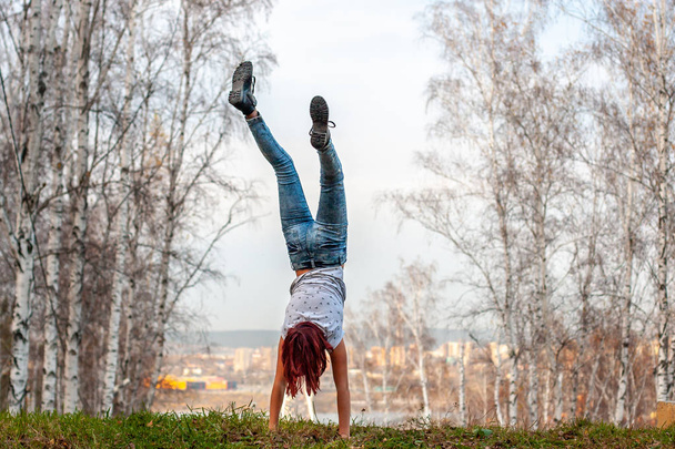 A girl with red hair stands on her hands with her back to the camera on a hill against the background of a blurred city and a birch forest. There is green grass on the ground. Acrobatics in nature. - Foto, Bild