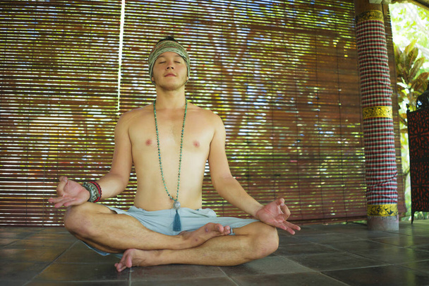 young handsome hipster meditation sitting in lotus position - Attractive athletic and happy man practicing yoga relaxation exercise wearing hair band and mala beads necklace - Photo, image