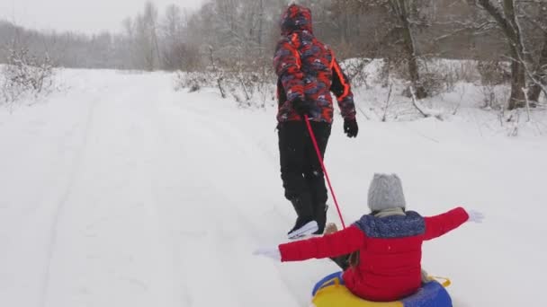 子供の雪の白い道に沿ってそりで幸せなお父さんは幸運です。クリスマス休暇と旅行。大人と子供のための楽しいゲーム。幸せな家庭の概念。十代の若者がチューブに乗る - 映像、動画