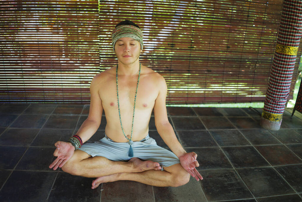 young handsome hipster meditation sitting in lotus position - Attractive athletic and happy man practicing yoga relaxation exercise wearing hair band and mala beads necklace - Foto, afbeelding