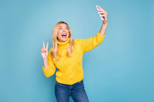 Photo of amazing blond lady holding telephone hands showing v-sign symbol making selfies wear knitted yellow pullover isolated blue color background - Photo, Image