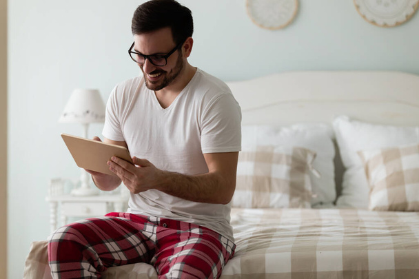 Όμορφος νεαρός χρησιμοποιώντας ψηφιακό tablet που κάθεται στο κρεβάτι στο σπίτι του - Φωτογραφία, εικόνα