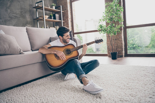 Photo of mixed race guy sitting floor leaning sofa hold acoustic instrumental guitar playing new written song sound signing creative talented person flat loft living room indoors - Fotó, kép
