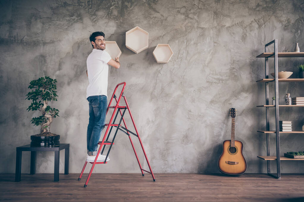 Full length profile view photo of mixed race guy installing wall shelf standing stepladder repair of new flat handmade working with pleasure hanging last shelf showing result in doors - Zdjęcie, obraz