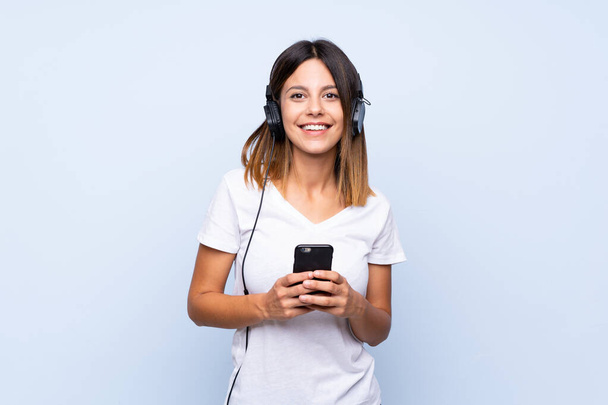 Young woman over isolated blue background using the mobile with headphones - Foto, immagini