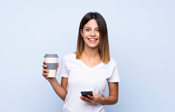 Молода жінка над ізольованим синім тлом тримає каву, щоб забрати і мобільний
 - Фото, зображення