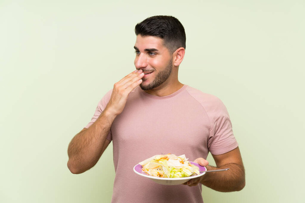 Jonge knappe man met salade over geïsoleerde groene muur die veel glimlacht - Foto, afbeelding