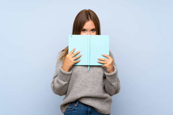 Έφηβος κορίτσι με πουλόβερ πάνω από απομονωμένο μπλε φόντο κρατώντας και διαβάζοντας ένα βιβλίο - Φωτογραφία, εικόνα