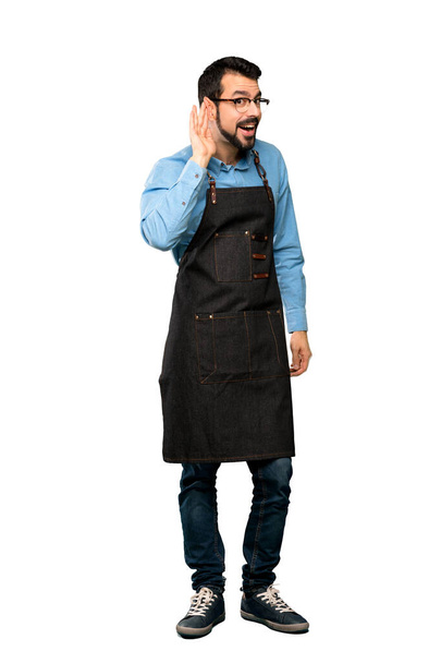 Full-length shot of Man with apron listening to something by putting hand on the ear over isolated white background - Foto, immagini