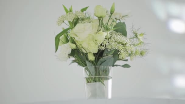 Pěkné bílé květy s velkými zelenými listy ve skleněné váze dát na bílý stůl. - Záběry, video