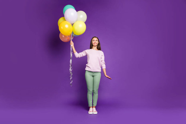 Full length body size photo of beautiful cheerful cute charming pretty sweet youngster looking pensively at air balls of helium wearing green pants trousers isolated over purple vivid color background - Fotoğraf, Görsel