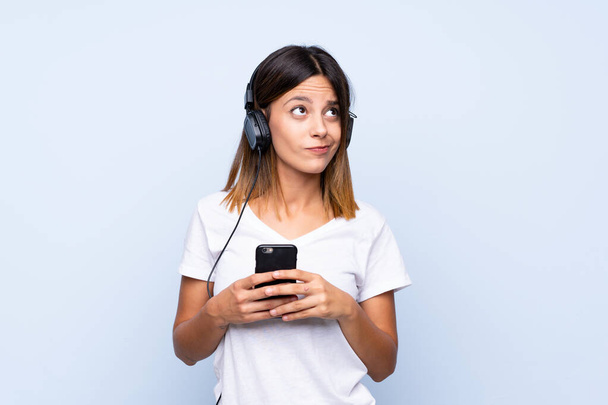 Mladá žena přes izolované modré pozadí pomocí mobilu se sluchátky a myšlení - Fotografie, Obrázek