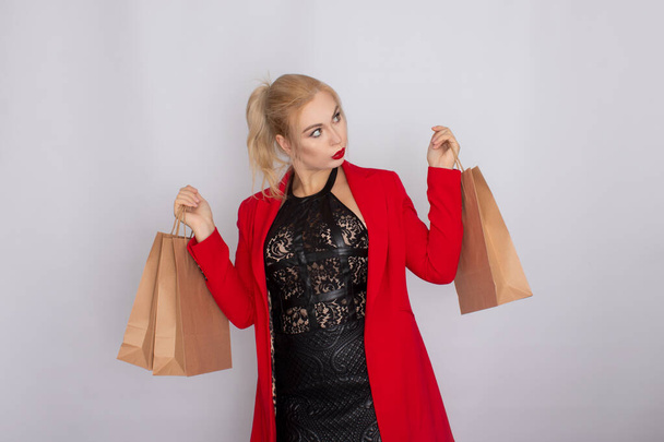 Image of excited blonde woman in red jacket holding brown shopping bags isolated on white bakcground - Фото, зображення