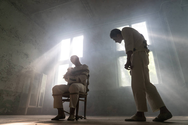 Un loco con camisa de fuerza está atado a una silla en una vieja clínica abandonada y el otro loco se acerca con interés.
 - Foto, Imagen