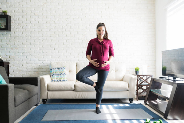 Muotokuva melko raskaana oleva nainen harjoitellaan jooga seisoessaan yhdellä jalalla olohuoneessa
 - Valokuva, kuva