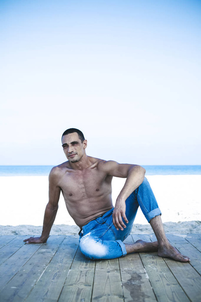 jonge gespierde man in denim broek rusten en poseren op het strand - Foto, afbeelding