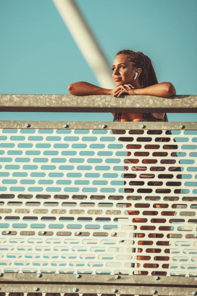 Nuori kunto nainen lepää rankan harjoittelun jälkeen joen sillalla, kuuntelee musiikkia ja katsoo pois
. - Valokuva, kuva