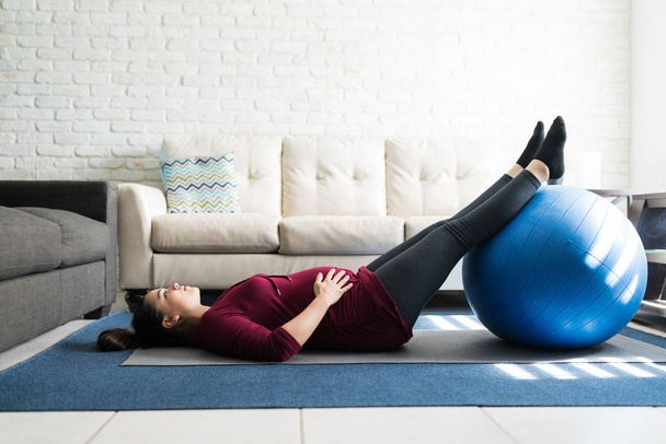 Full length of good looking pregnant female lying on exercise mat with ball by sofa at home - Photo, Image