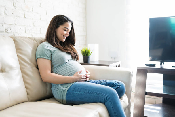 Beautiful pregnant female touching while admiring her baby bump and smiling at home - Foto, Bild