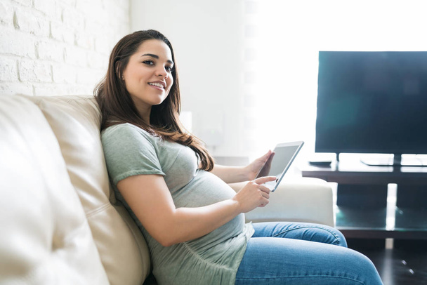 attraktive Schwangere mit digitalem Tablet, während sie Blickkontakt aufnimmt und zu Hause lächelt - Foto, Bild