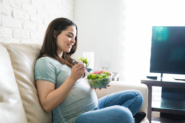 Gyönyörű várandós nő eszik friss egészséges salátát tálból, miközben otthon ül - Fotó, kép