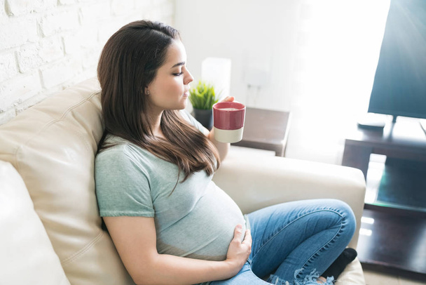 Привлекательная беременная женщина расслабляется, выпивая свежий кофе из кружки и расслабляясь дома
 - Фото, изображение