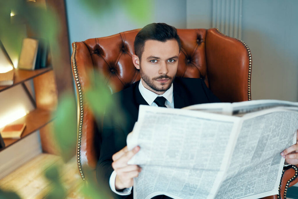 Zelfverzekerde jonge zakenman leest krant en het laatste nieuws terwijl hij in de fauteuil zit  - Foto, afbeelding