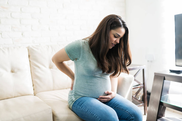Mooie zwangere vrouw die lijdt aan rugpijn tijdens het zitten op de bank tijdens haar zwangerschap in de woonkamer - Foto, afbeelding