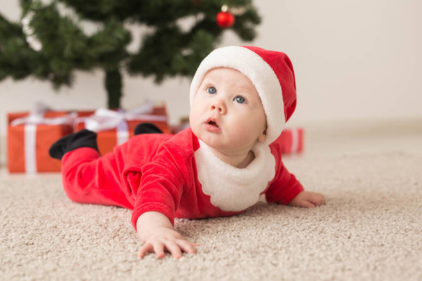 Милая малышка в костюме Санта Клауса ползает по полу над елкой. Праздничный сезон
. - Фото, изображение