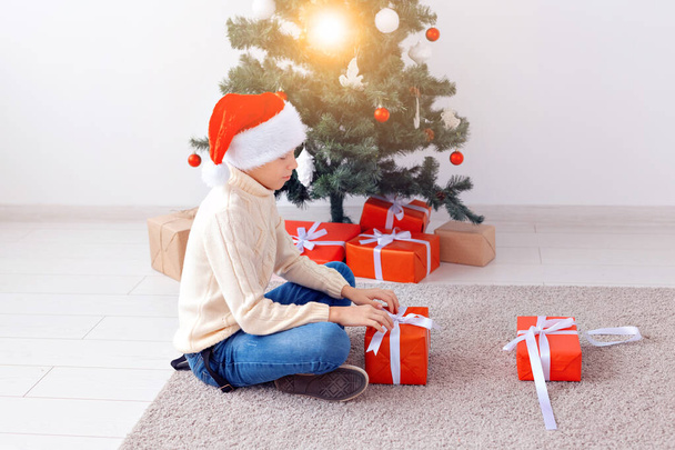 Свята, Різдво, дитинство та концепція людей усміхнений щасливий хлопчик-підліток у капелюсі Санти відкриває подарункову коробку на фоні ялинки
 - Фото, зображення
