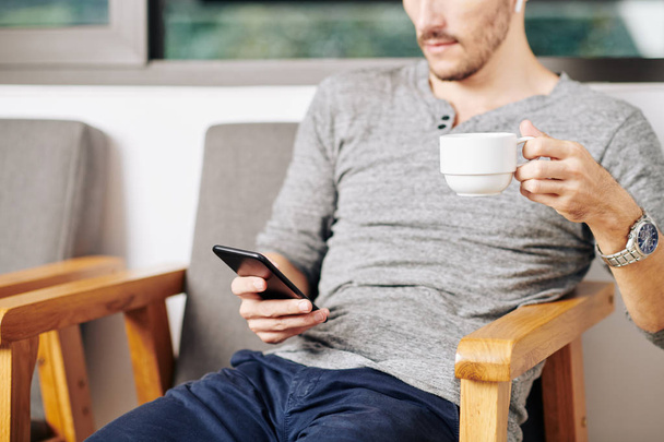 Imagen recortada del hombre descansando en el sillón, bebiendo taza de té y leyendo noticias en el teléfono inteligente
 - Foto, imagen