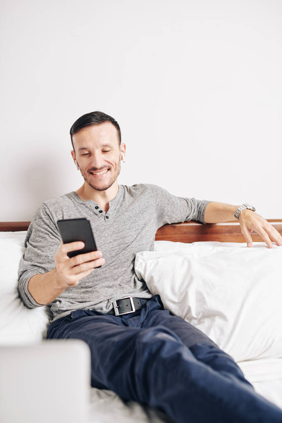 lächelnder fröhlicher junger Mann, der auf dem Bett sitzt, wenn er lustige Nachrichten von Freunden auf dem Smartphone liest - Foto, Bild