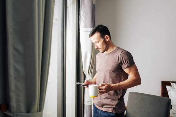 Pensive nuori mies älypuhelin juo aamukahvia ja tarkistaa sosiaalisen median
 - Valokuva, kuva
