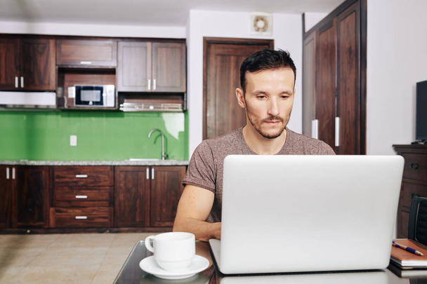Серьезный студент университета сидит за столом в своей студии и работает на ноутбуке
 - Фото, изображение