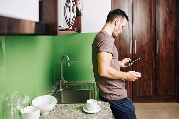 Чоловік спирається на кухонний лічильник і використовує мобільний додаток для замовлення доставки їжі
 - Фото, зображення