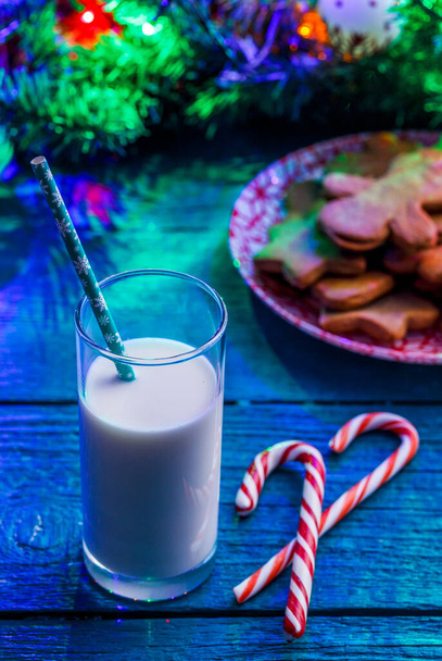 Picture of blue table with Christmas cookies, glass of milk, caramel sticks, spruce branches with burning garland - Fotografie, Obrázek
