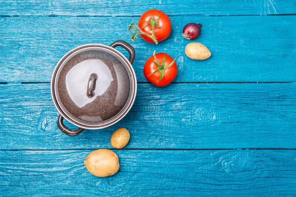 Photo of iron pot, tomato, potato, onion on blue wooden background. Empty space for text - Foto, Bild