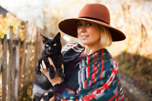 Black cat and stylish traveling woman wearing  authentic boho chic style poncho and hat near old wooden countryside buildings Woman have a fun in autumn time.Travel and wanderlust concept. - Foto, imagen