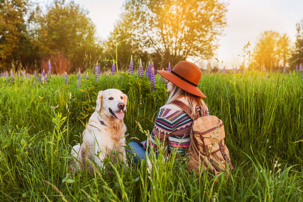 Traveling woman and golden retriever dog in nature. Stylish hipster in brown hat and boho style sweater with vintage textile backpack in summer green meadow.  - Fotó, kép