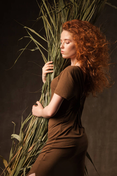 Energizer beautiful young curly ginger hair woman with green tropical plant cane grass posed in dark walls studio.  - Photo, image